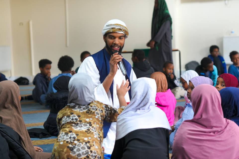 A teacher with a microphone among children seated on the carpet