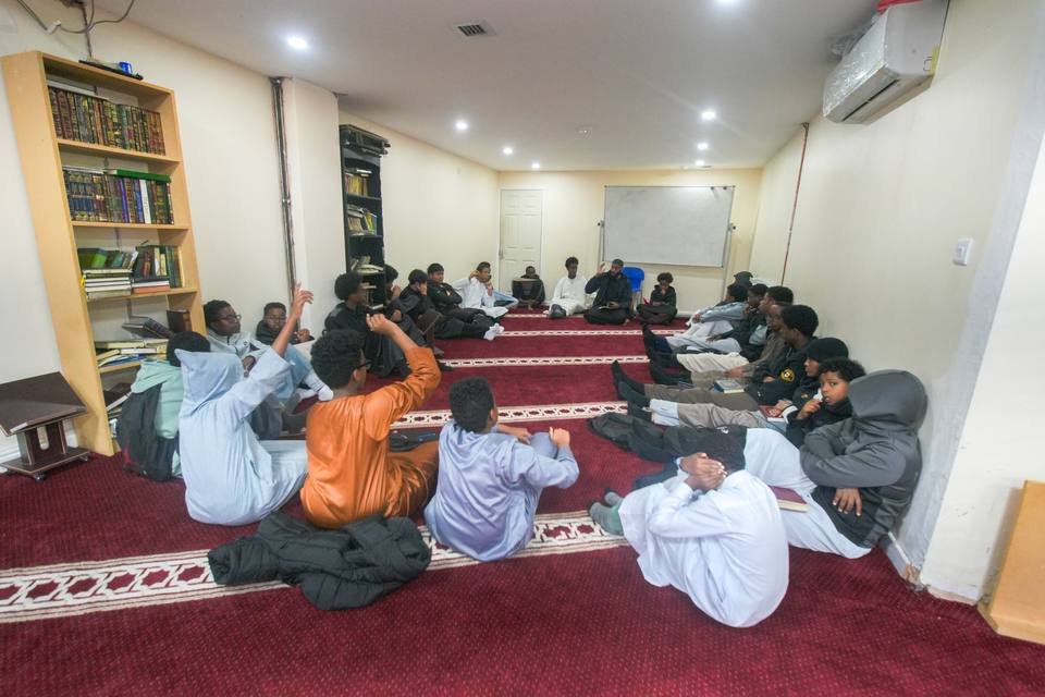 A class of boys sitting round the room on the red carpet, their teacher at the far end
