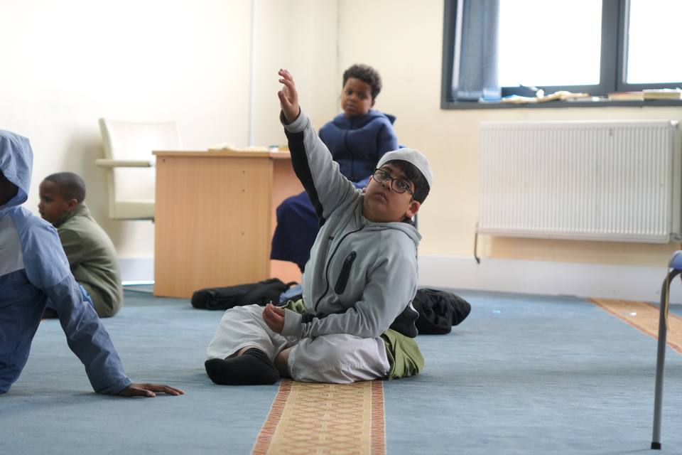 A boy sitting on the carpet with his hand raised