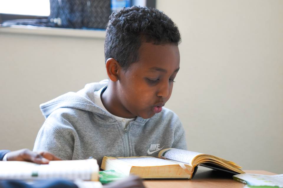 A boy reading aloud from a book open on the table