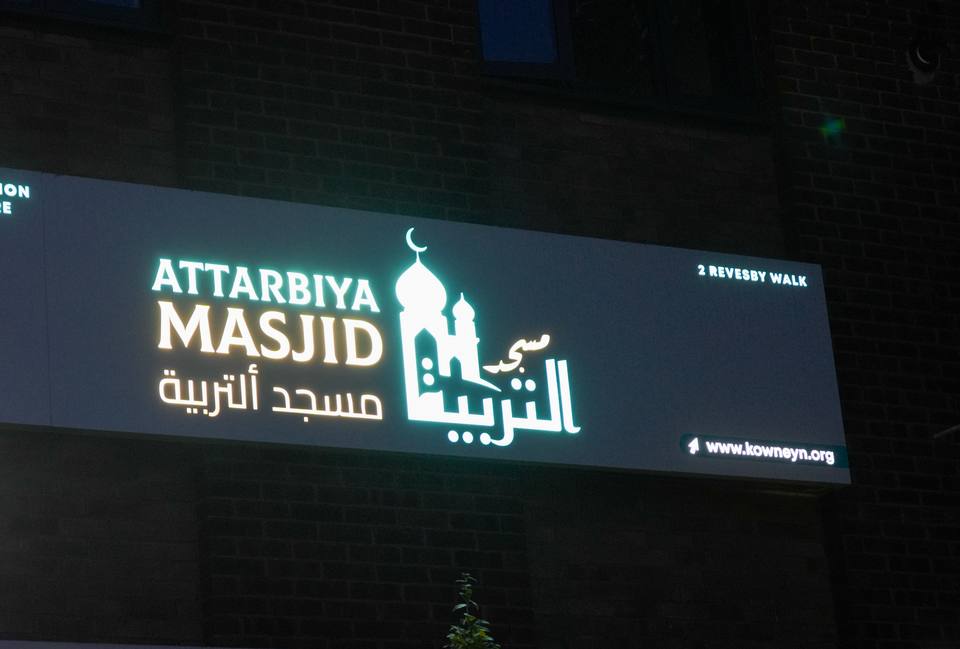 The masjid's sign lit at night: Attarbiya Masjid in English and Arabic