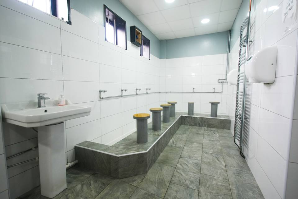 The wudu area: white tiles from floor to ceiling, taps along the wall and a low seat at each