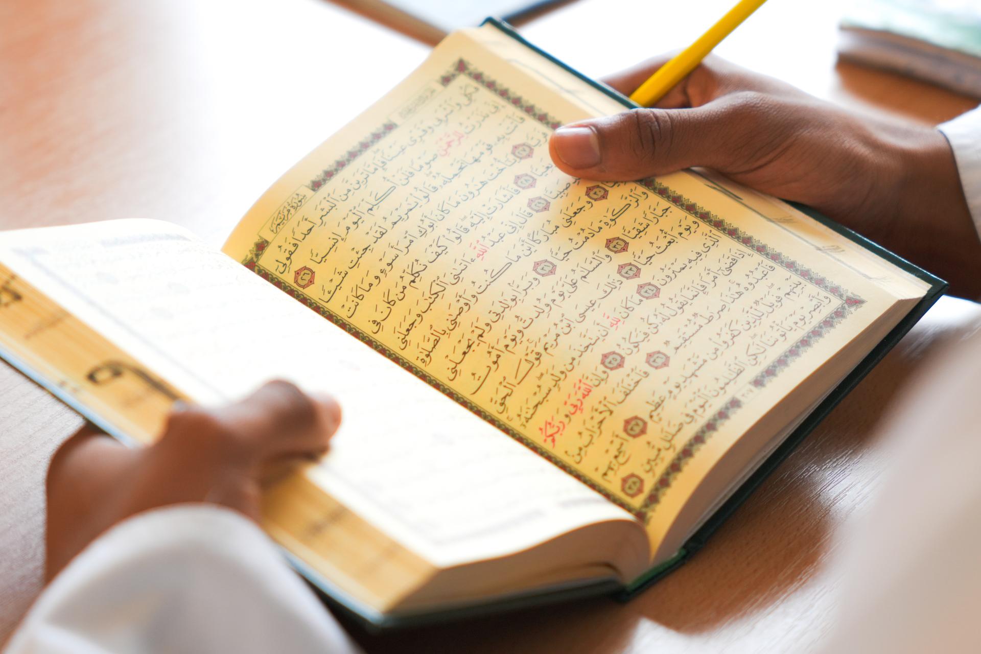 A pupil's hands holding an open Qur'an, a pencil resting on the page