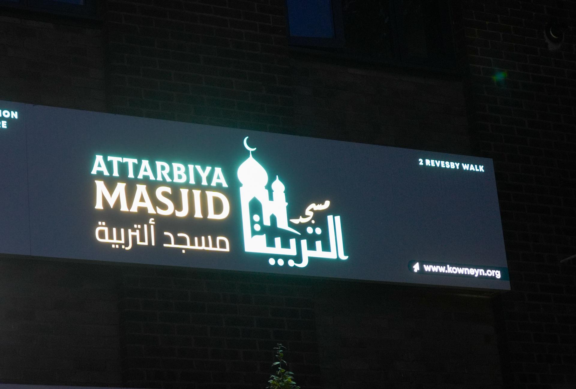 The masjid's sign lit at night: Attarbiya Masjid in English and Arabic