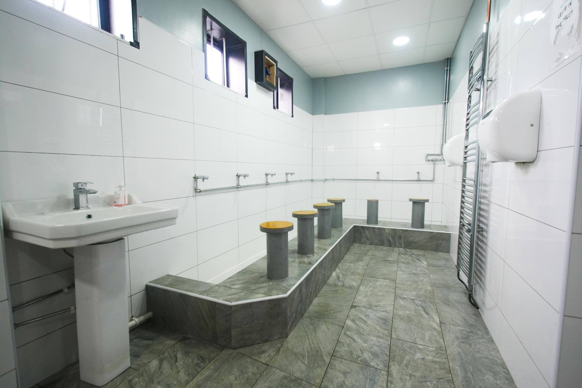 The wudu area: white tiles from floor to ceiling, taps along the wall and a low seat at each